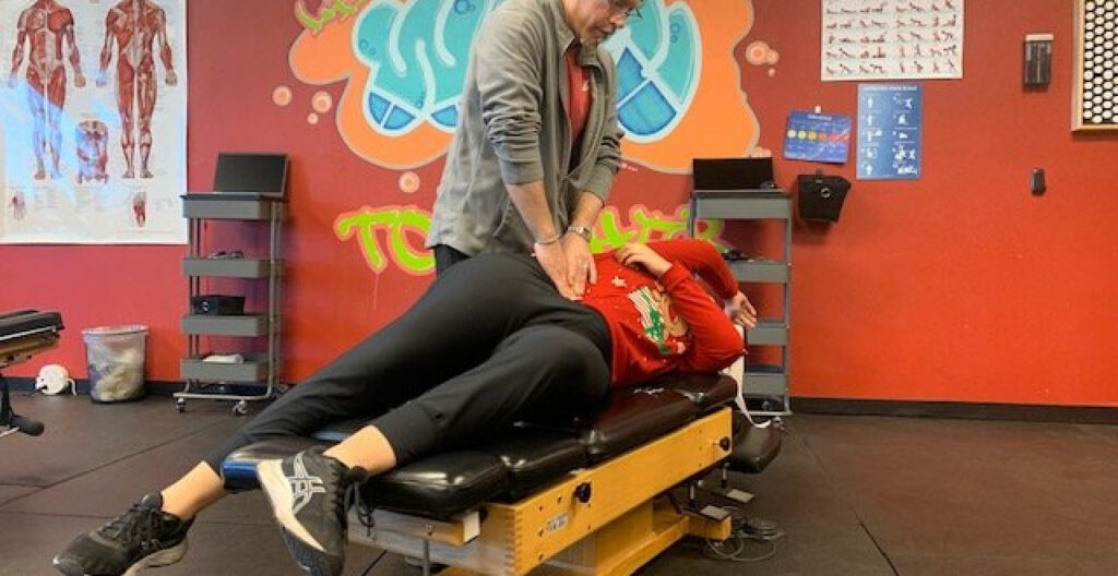 A person lies on their side on a chiropractic table while a practitioner, known for treating sports injury Nevada cases, performs a spinal adjustment in a clinic room adorned with anatomical posters.
